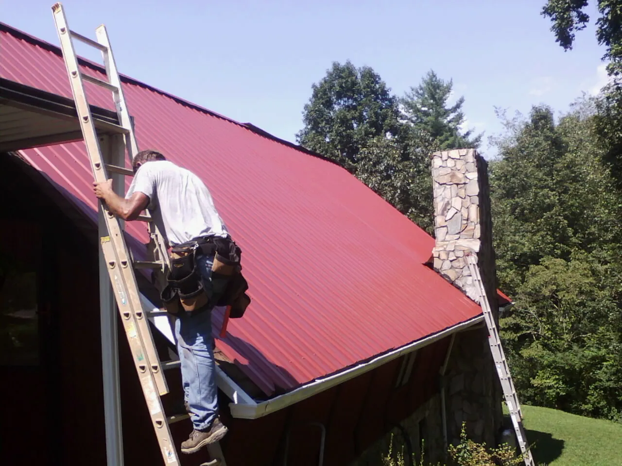 Roofer on a ladder beside a red metal roof and stone chimney — Soffit & Fascia Repair by Roofing Brothers in Hollister
